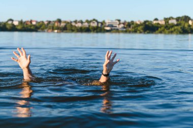 Gölde boğulan biri yardım için çaresizce ellerini kaldırıyor. Arka planda bulanık bir şehir silüeti beliriyor.