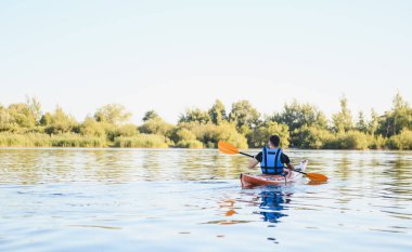 Kayakçı, yemyeşil yemyeşil bir göl üzerinde, izleyiciden uzaklaşıyor, açık havada huzurlu bir dinlenme anının tadını çıkarıyor.