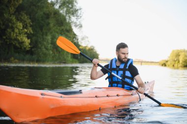 Yazın nehirde turuncu kano süren bir adam can yeleği giyiyor ve çift kanatlı kürek kullanıyor.