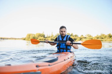 Güneşli bir günde turuncu bir kanoyla nehirde kürek çeken, can yeleği giyen ve kürek çeken, açık havada maceranın tadını çıkaran bir adam.