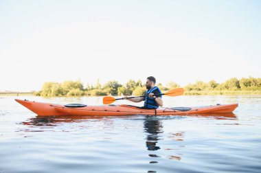 Can yeleği giyen bir adam sakin bir gölde ya da nehirde turuncu bir kanoyla kürek çeker. Güneşli bir günde su sporlarının ve aktif eğlencelerin tadını çıkarır.