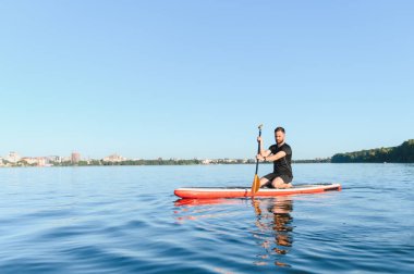 Genç sporcu yemek masasında kürek çekiyor, gölde güneşli bir günün tadını çıkarıyor, arka planda şehir manzarası var.