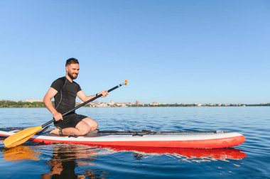 Genç sporcu, arka planda güneşli bir yaz gününün tadını çıkarırken sakin bir gölde yemek masasında diz çökerek kürek çekiyor.