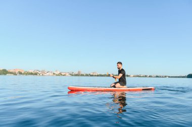 Genç sporcu, yemek masasında diz çökmüş, ufukta şehir manzarası olan bir gölde güneşli bir günün tadını çıkarıyor.