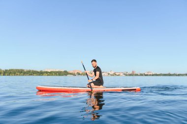 Genç sporcu arka planda manzarası olan sakin bir gölde yemek masasında diz çökerken kürek çekiyor.