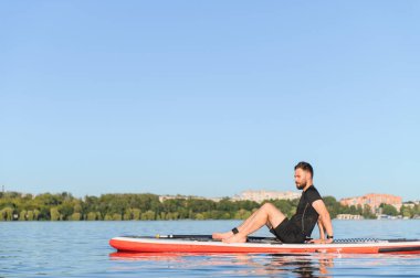 Genç sporcu, güneşli mavi gökyüzünün altındaki dingin bir gölde canlandırıcı bir antrenmandan sonra ayakta duran bir kürek tahtasında dinleniyor.