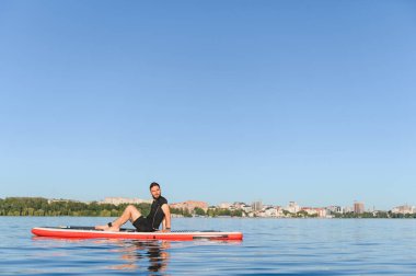 Huzur dolu bir anın tadını çıkaran genç bir adam durgun bir nehirde, arka planda şehir silüeti olan kürek tahtasında oturuyor.