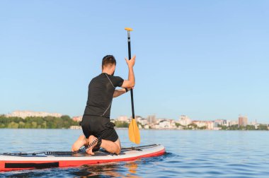 Ayakta duran bir sörf tahtasına diz çöken, ufukta görünen bir şehir manzarasıyla sakin bir gölde kürek çeken sporcuların arka görüntüsü.