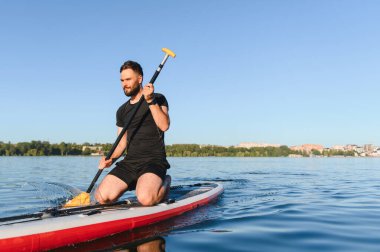 Genç sporcu ayakta kürek çekiyor, güneşi emiyor ve sakin göl sularında güzel bir günün tadını çıkarıyor.