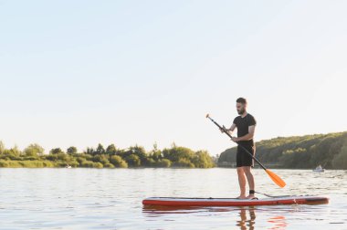 Genç adam gün batımında bir gölün üzerinde ayakta kürek çekiyor, su sporunun ve aktif eğlencelerin tadını çıkarıyor.