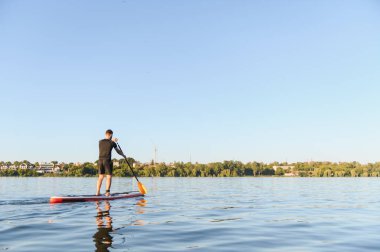 Sporcu, sakin bir gölün üzerinde suyla kürek çekiyor. Açık mavi gökyüzü ve uzak bir şehir manzarasıyla yaz gününün tadını çıkarıyor.