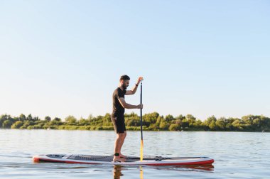 Genç sporcu, gün batımında sakin bir gölde, su sporu ve aktif eğlence için yemek masasında kürek çekiyor.