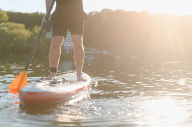 Gün batımında ayakta kürek çeken bir adam gölün huzurlu sükunetinde altın saat ışığı suyun üzerinde dans ederken sırılsıklam oluyor.