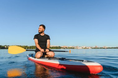Genç sporcu, açık mavi gökyüzünün altındaki dingin göl manzarasının tadını çıkarırken elinde kürekle yemek masasında oturuyor.