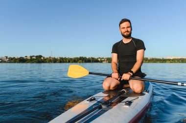 Genç sporcu, bir gölde ayakta duran bir sörf tahtasında diz çökerken, yaz gününün tadını çıkarıyor.