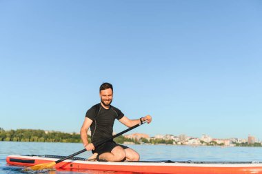 Sakallı adam yemek masasında diz çökerken, şehrin yakınındaki bir gölde yaz gününün tadını çıkarıyor.