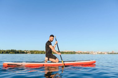 Genç sporcu gölün üzerinde bir yemek masasında kürek çekiyor, şehrin önünde güneşli bir günün tadını çıkarıyor.