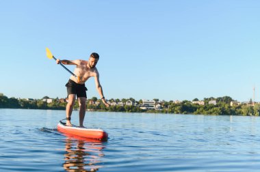 Genç adam durgun bir gölde güzel bir yaz gününün tadını çıkarırken ayakta kürek çekiyor ve dengesini koruyor.