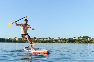 Genç atletik bir adam yemek masasında dengede duruyor, elinde bir kürek tutuyor, su sporlarının ve gölde aktif olarak eğlenmenin tadını çıkarıyor.