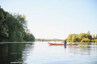 Kanocu, gün batımında yemyeşil yemyeşil ve uzak bir köprüyle çevrili sakin bir nehirde huzurlu bir kürek çekiyor.