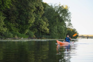 Güneşli yaz gününde yeşil ormanın yakınında sakin bir nehirde kayak yapan bir adam su sporu yapıyor.