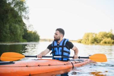 Genç adam güneşli bir günde nehirde kayak yapıyor, can yeleği giyiyor ve turuncu raketle kürek çekiyor, aktif eğlence ve su sporlarının tadını çıkarıyor.