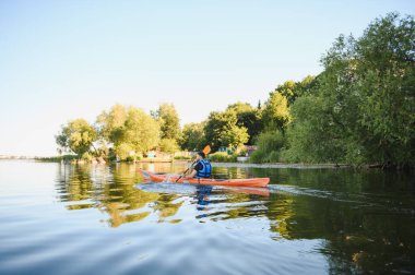Bereketli yeşil ağaçlarla çevrili sakin gölde kürek çeken bir adam, güneşli bir su sporları ve aktif dinlenme gününün tadını çıkarıyor.