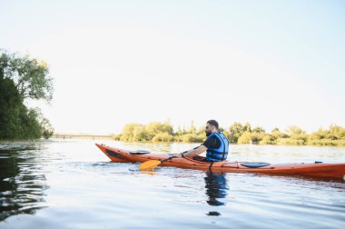 Gün batımında sakin bir nehirde kano süren bir adam, doğanın huzurunun tadını çıkarıyor ve aktif olarak dinleniyor.