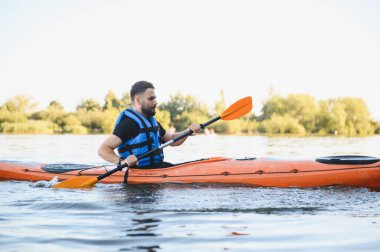 Sakin bir gölde turuncu bir kanoyla kürek çeken bir adam, yaz günü su sporlarının ve aktif bir dinlenmenin tadını çıkarıyor.