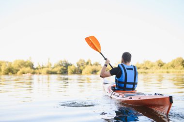 Can yelekli bir adam sakin bir gölde turuncu bir kanoyla kürek çekiyor, güneşli bir günde su sporları ve aktif eğlence yapıyor.