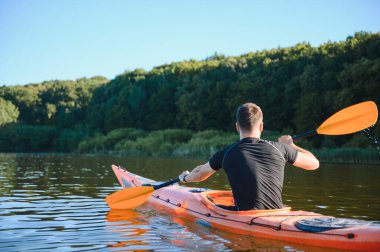 Arka planda yemyeşil bir ormanla sakin bir gölde kürek çeken kanocu, doğada huzurlu bir anın tadını çıkarıyor.