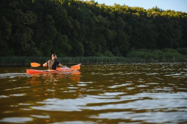 Güzel bir günbatımında yeşil bir ormanın yakınındaki sakin bir gölde kürek çeken kanocu, doğanın huzurunun tadını çıkarıyor.