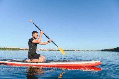 Genç sporcu ayakta duran bir sörf tahtasına diz çöküp güneşli bir yaz gününde sakin bir gölde kürek çekiyor.