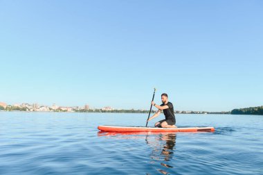 Güneşli bir günde arka planda şehir manzarası olan bir gölde kürek çekme alıştırması yapan genç adam.