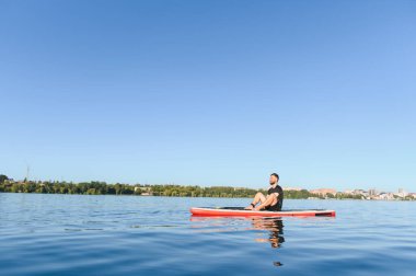 Genç adam ayakta bir tahtanın üzerinde oturuyor ve dinleniyor güneşli bir yaz gününde gölün sessizliğinin tadını çıkarıyor.