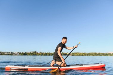 Genç sporcu, gölde güneşli bir yaz gününün tadını çıkarırken yemek masasında diz çökerek kürek çekiyor.