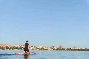 Ayakta duran bir adam arka planda şehir manzarasıyla sakin sularda süzülür, aktif eğlence ve su sporlarını gözler önüne serer.