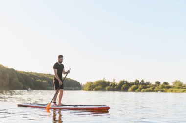 Genç sporcu ayakta kürek çekiyor, güneşli bir yaz günü doğanın güzellikleriyle çevrili sakin bir gölde eğleniyor.