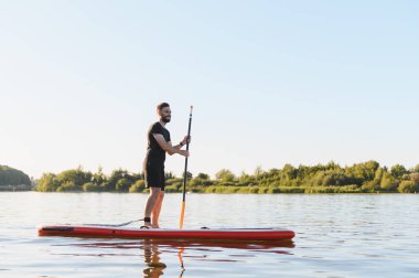 Genç bir sporcu yemek masasında kürek çekiyor, güneş batarken gölde geçireceği bir yaz akşamının tadını çıkarıyor, huzurlu bir atmosfer yaratıyor.