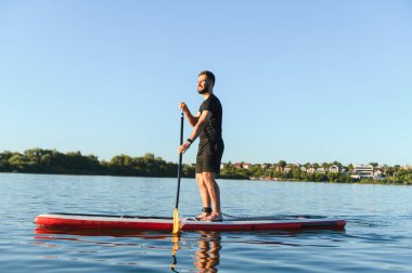 Genç sporcu göl kenarında bir yemek masasında kürek çekiyor, bir yaz günü su sporu aktivitesinin tadını çıkarıyor.