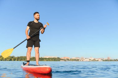 Genç sporcu arka planda su sporlarının tadını çıkaran bir şehir manzarası olan göldeki bir panoda kürek çekiyor.