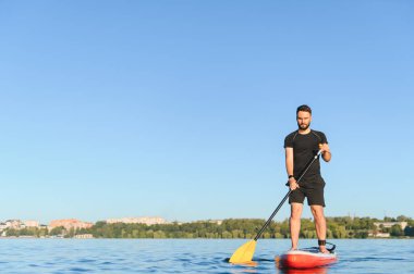 Genç sporcu gölün üzerinde bir panoda kürek çekiyor, ayakta sörf yapıyor, su sporlarının ve yaz aktivitelerinin tadını çıkarıyor.
