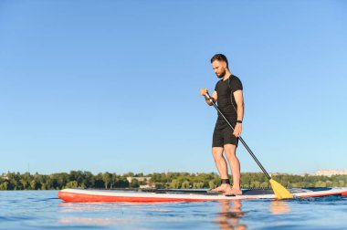 Genç sporcu, durgun bir gölün üzerinde durmaktan zevk alıyor açık mavi bir gökyüzünün altında, sağlıklı ve aktif bir yaşam tarzını destekliyor.