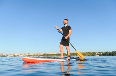 Genç sporcu, kentteki bir gölde ayakta kürek çekiyor. Su sporunun ve aktif eğlencelerin tadını çıkarıyor.