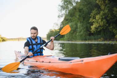Sahil kenarındaki sakin bir nehirde turuncu bir kanoya binen adam, su sporlarının ve doğada aktif olarak eğlenmenin tadını çıkarıyor.