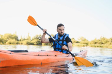 Nehirde veya gölde turuncu kano süren bir adam can yeleği giyiyor, su sporunun ve doğada aktif eğlencenin tadını çıkarıyor.