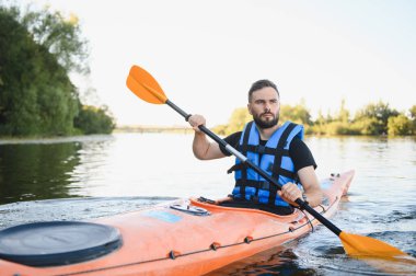 Adam güneşli bir nehirde turuncu bir kanoya biniyor, su sporlarının heyecanını yaşıyor ve aktif bir açık hava yaşam tarzını kucaklıyor.