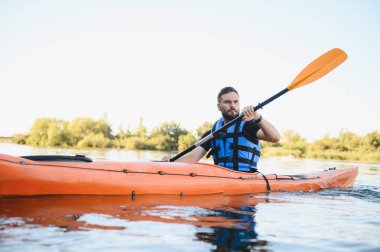 Can yeleği giymiş bir adam sakin suda turuncu kano sürüyor. Aktif eğlence ve su sporlarının tadını çıkarıyor.