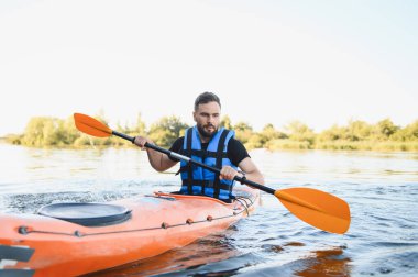 Nehirde veya gölde turuncu kano süren bir adam can yeleği giyiyor, yaz günü su sporlarının keyfini çıkarıyor.