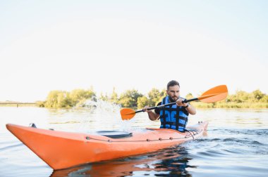Can yeleği giyen bir adam nehirde turuncu bir kanoya kürek çeker ve küreğiyle su sıçratır.